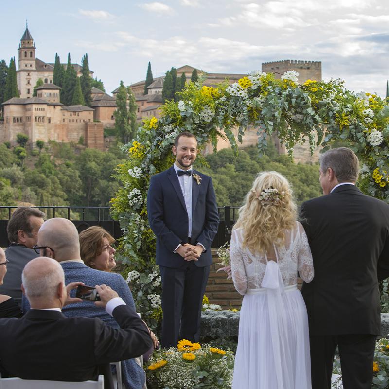 bodas-elcamborio-granada-galeria-10 Novios durante ceremonia civil exterior con la alhambra de granada de fondo y decoración floral